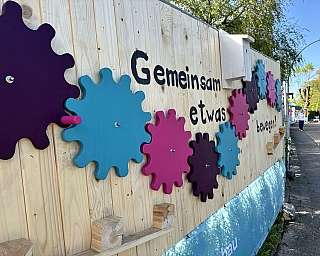 Holzwand mit bunten Zahnrädern und Text in einem Außenbereich an einem Gehweg